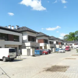 Ulica z szeregiem nowych, dwukondygnacyjnych domów w zabudowie szeregowej z garażami, wykończonych w bieli i brązie, z zaparkowanymi samochodami i kontenerami na śmieci.