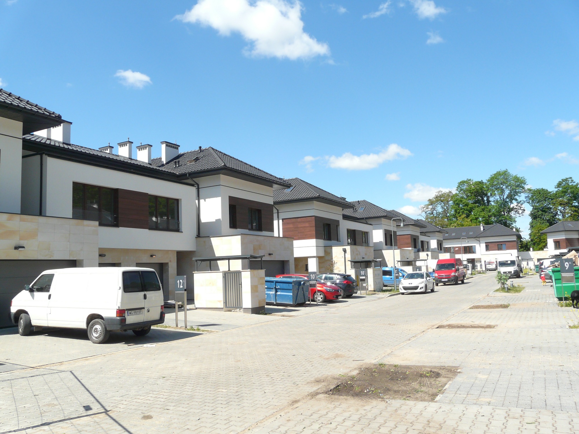 Ulica z szeregiem nowych, dwukondygnacyjnych domów w zabudowie szeregowej z garażami, wykończonych w bieli i brązie, z zaparkowanymi samochodami i kontenerami na śmieci.