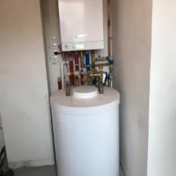 Biały zasobnik ciepłej wody użytkowej z piecem gazowym Viessmann zamontowany na ścianie w surowym pomieszczeniu z widocznymi instalacjami hydraulicznymi.