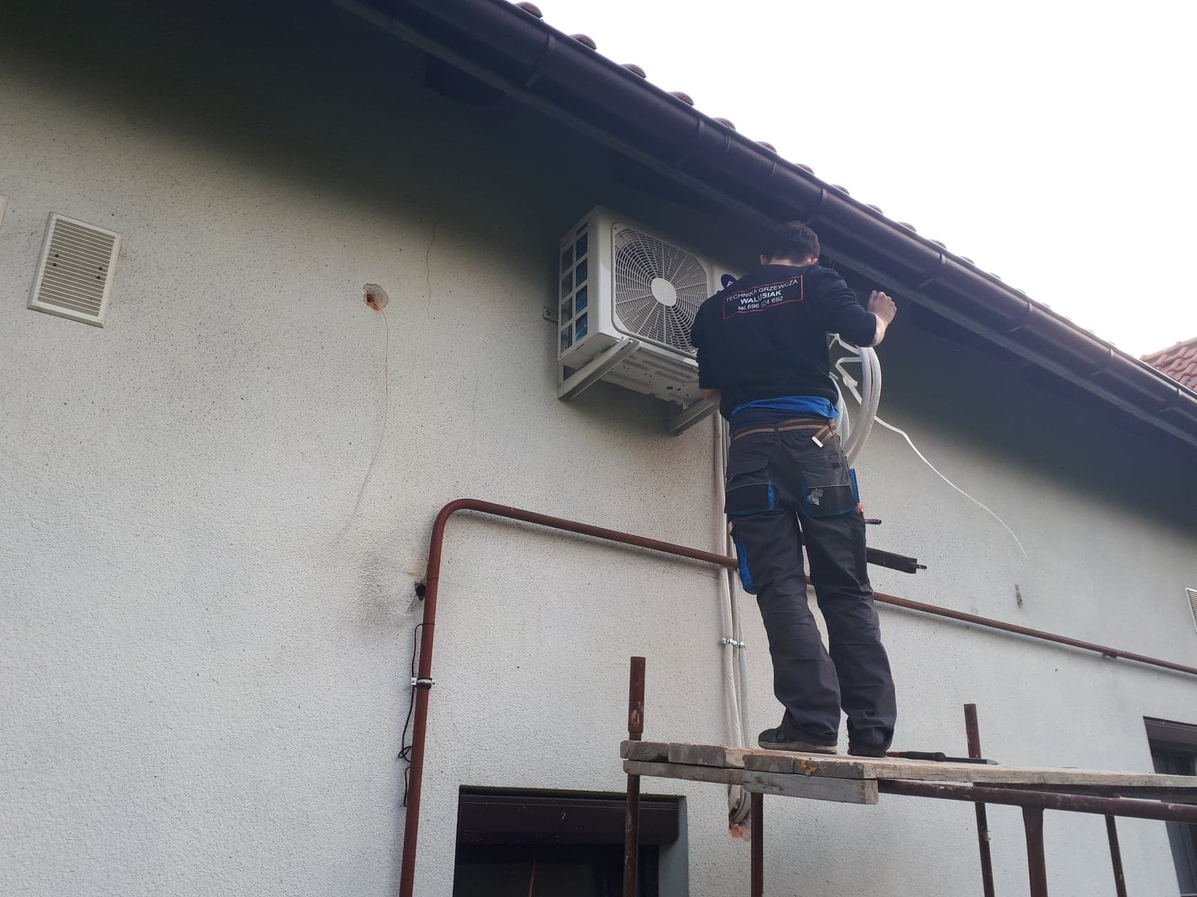 Instalator na rusztowaniu montuje zewnętrzną jednostkę klimatyzacji na elewacji budynku, widoczny fragment rynny i rura instalacyjna na ścianie.