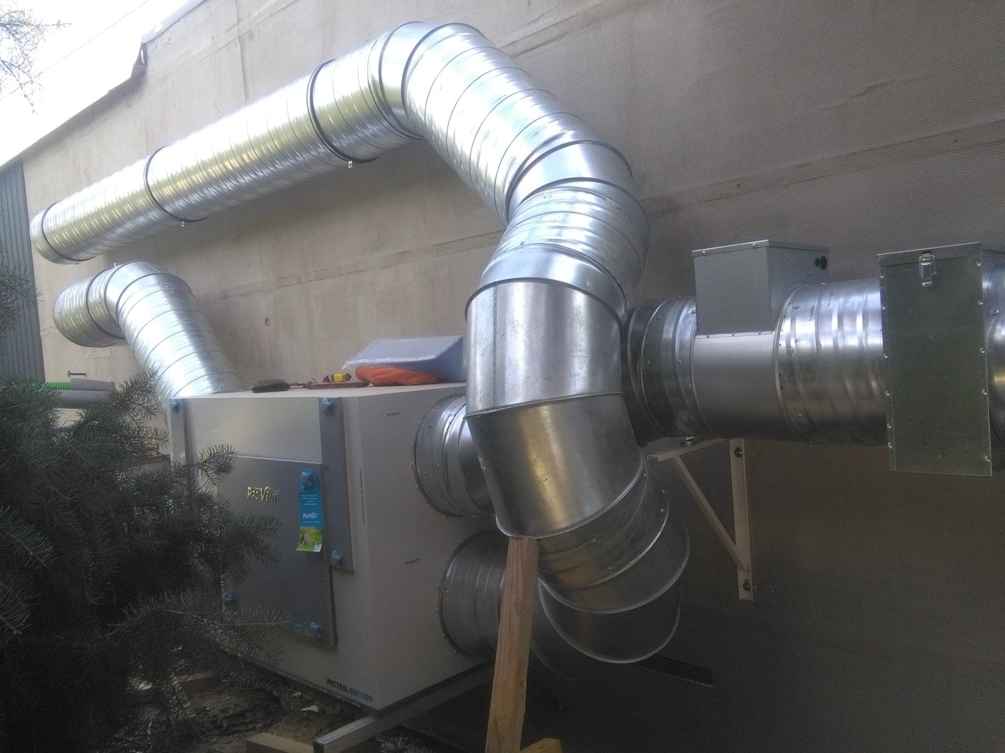 Centrala wentylacyjna z rekuperacją ProVent Mistral BSR 1100 z podłączonymi, srebrnymi kanałami wentylacyjnymi na zewnątrz budynku, widoczne narzędzia i akcesoria montażowe.