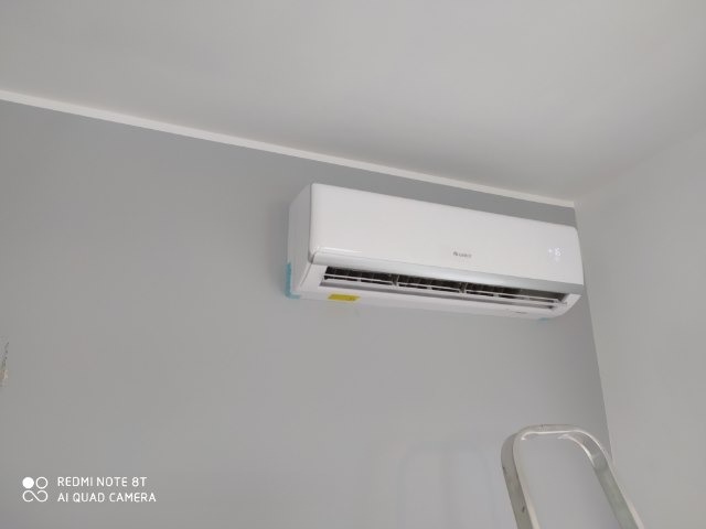 Biały klimatyzator marki Gree zamontowany na szarej ścianie, widoczny fragment drabiny malarskiej, minimalistyczne wnętrze.
