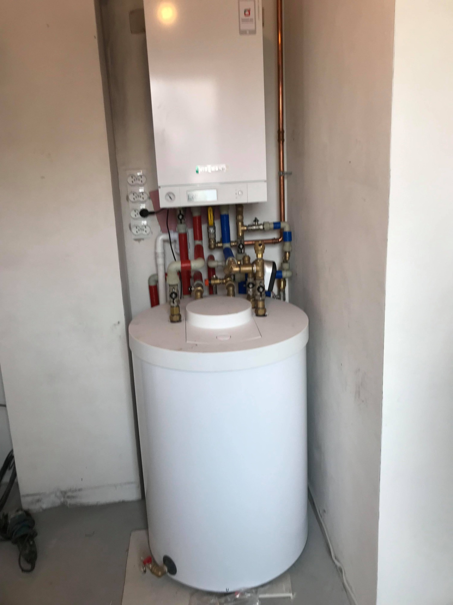 Biały zasobnik ciepłej wody użytkowej z piecem gazowym Viessmann zamontowany na ścianie w surowym pomieszczeniu z widocznymi instalacjami hydraulicznymi.