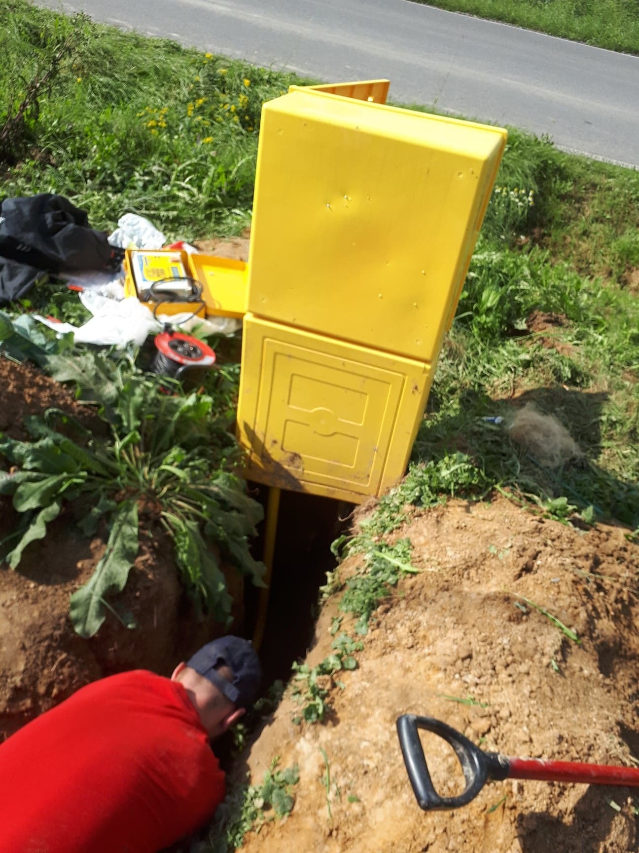 Instalacja przyłącza gazowego: żółta skrzynka gazowa wystająca z wykopu, widoczny fragment rury, obok leżą narzędzia i odzież robocza, a pracownik w czerwonej koszulce pochyla się nad wykopem.