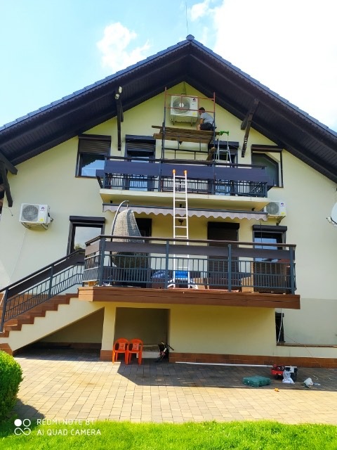 Montaż klimatyzacji na elewacji domu jednorodzinnego: pracownik na rusztowaniu przy jednostce zewnętrznej, widoczna drabina oparta o balkon, narzędzia na ziemi.