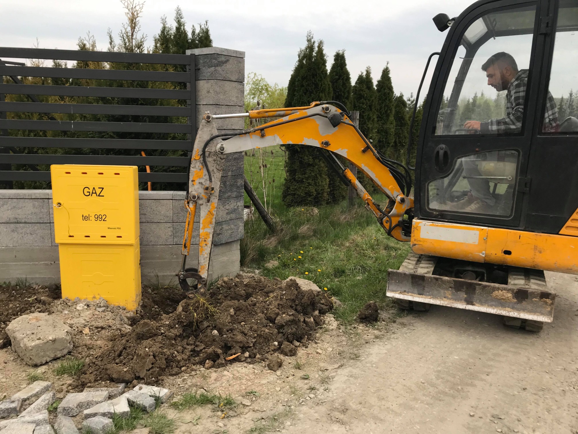 Żółta skrzynka gazowa obok wykopu wykonanego koparką, w tle szary płot i tuje.