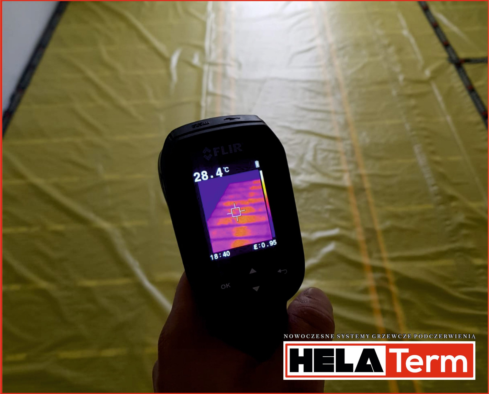 Termowizja podczerwieni ukazuje rozkład temperatury na folii grzewczej podłogowej, trzymana w dłoni, na ekranie temperatura 28.4°C.