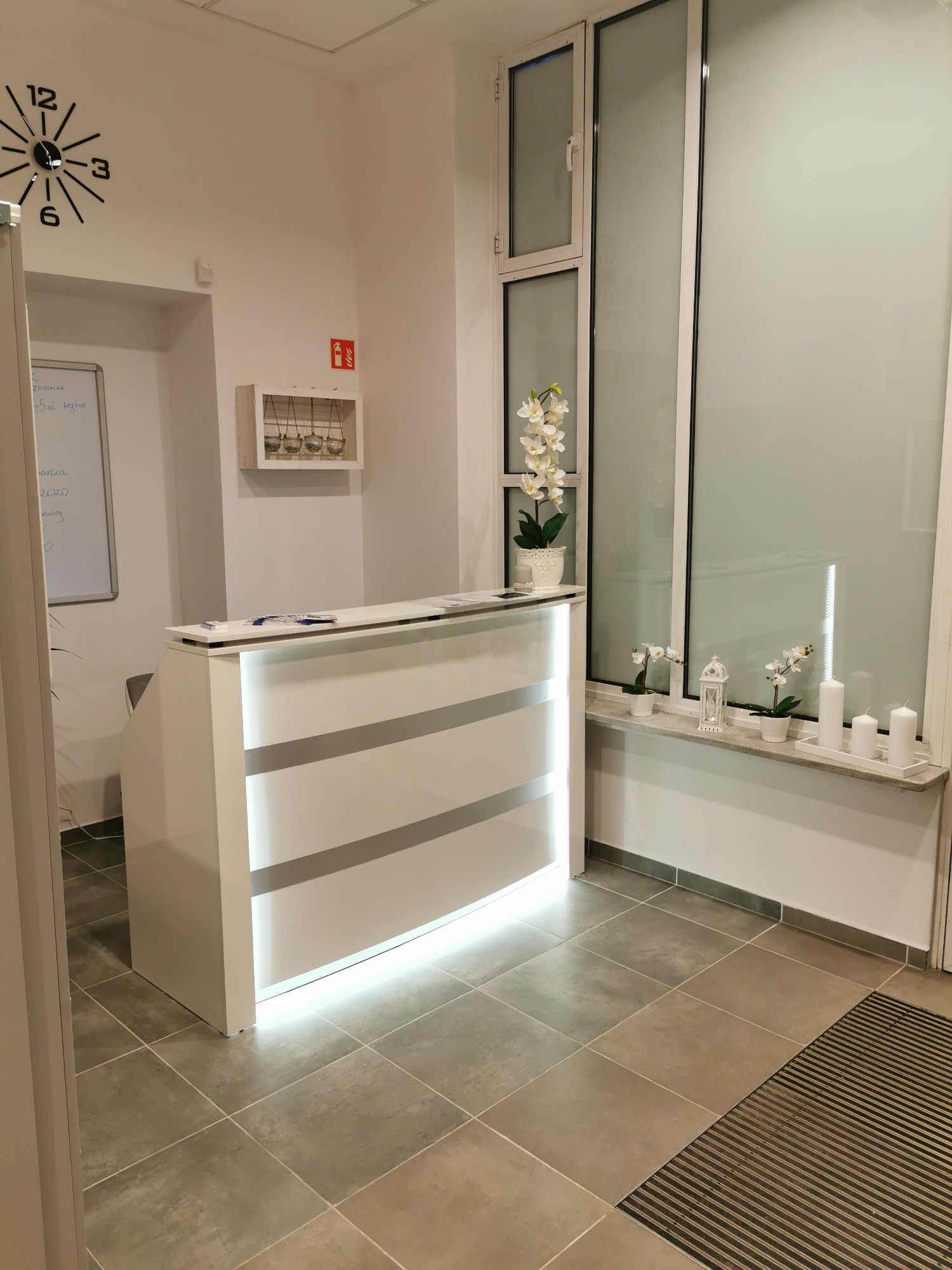 Jasne, nowocześnie urządzone wnętrze recepcji z białym biurkiem podświetlanym od dołu, kwiatami w doniczkach na parapecie i zegarem na ścianie.