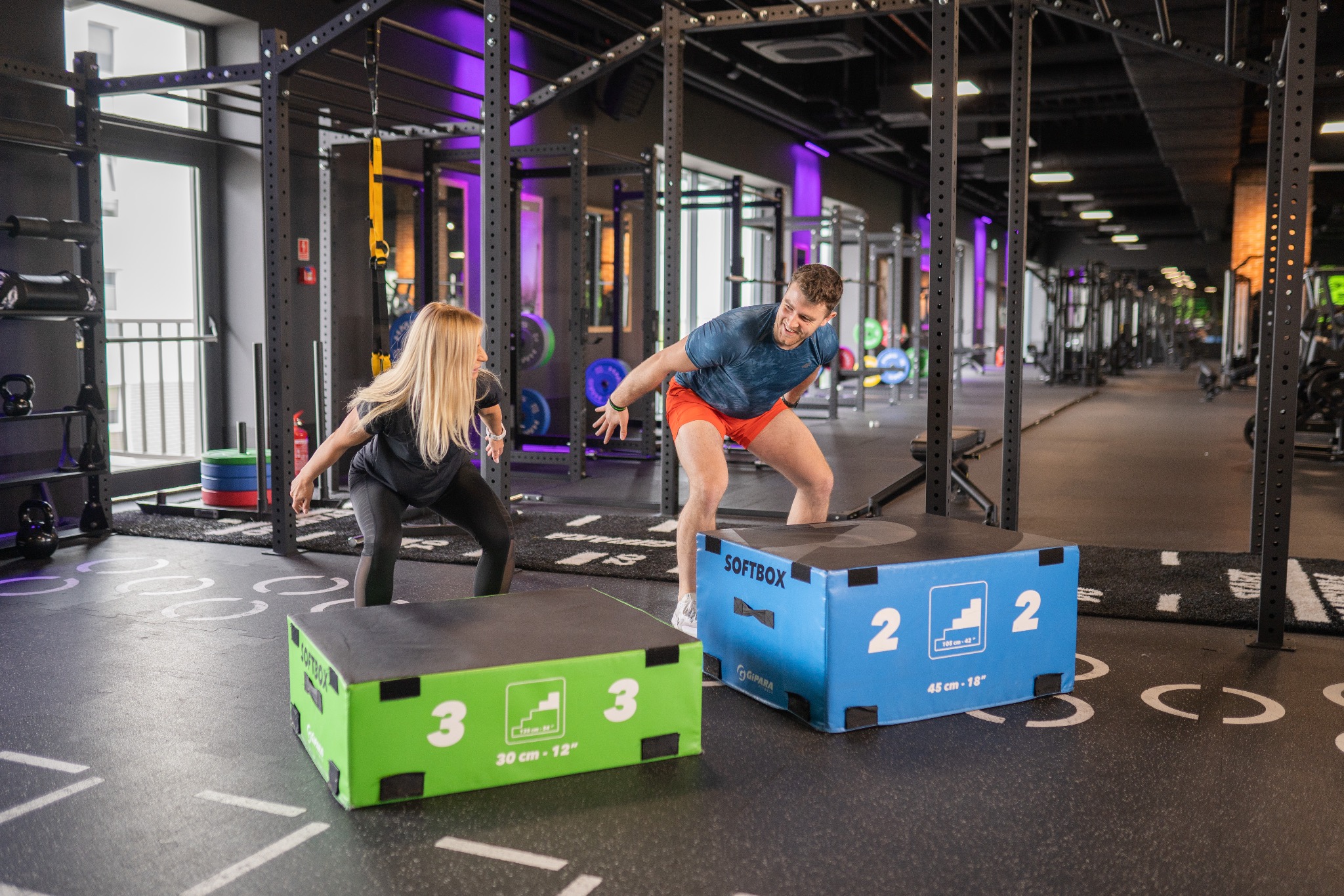 Kobieta i mężczyzna przygotowują się do skoku na skrzynie w nowoczesnej siłowni z fioletowym oświetleniem akcentującym, widoczne skrzynie do ćwiczeń Softbox w kolorach zielonym i niebieskim.