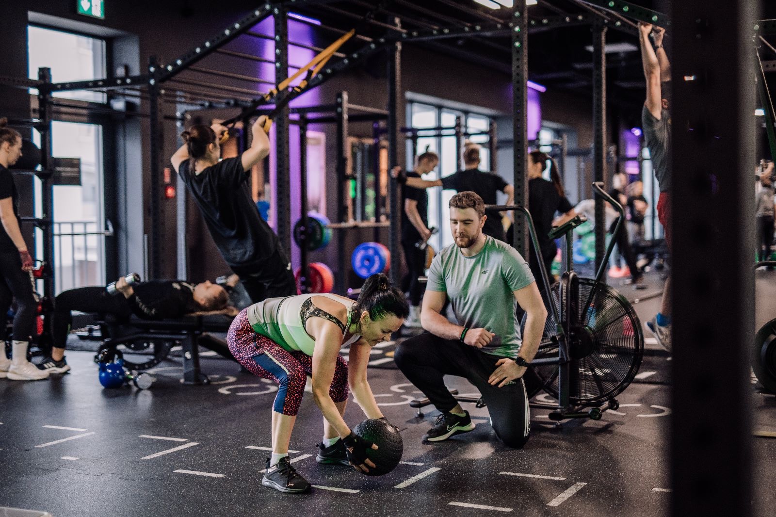 Kobieta w sportowym stroju podnosi piłkę lekarską pod okiem trenera personalnego w nowoczesnej siłowni z innymi ćwiczącymi w tle.
