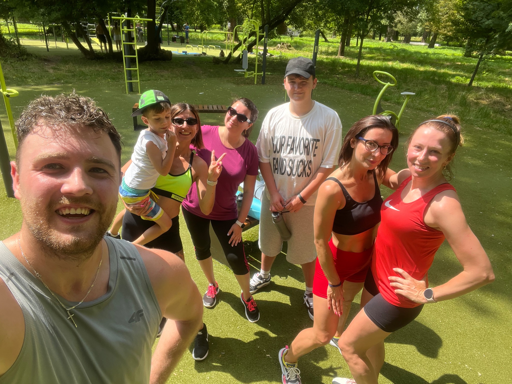 Grupa uśmiechniętych osób, w tym dziecko, pozuje do selfie na tle plenerowej siłowni z zieloną nawierzchnią i drzewami. Widoczny sprzęt do ćwiczeń.