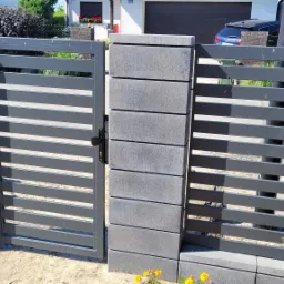 Nowoczesna brama wjazdowa z poziomymi, szarymi przęsłami, osadzona na słupkach z szarej kostki betonowej, z widocznym zamkiem i fragmentem ogrodzenia w tle.