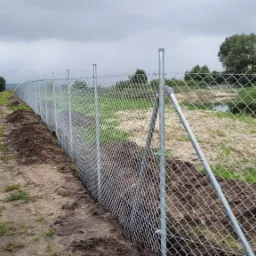 Długa siatka ogrodzeniowa, widok z boku, z podporami ukośnymi, rozciągnięta wzdłuż polnej drogi, za ogrodzeniem widać rzekę i drzewa, pochmurne niebo.