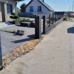 Montaż ogrodzenia betonowego z czarnych bloczków, widoczny etap budowy słupków i podmurówki, częściowo ułożona kostka brukowa, zaparkowany samochód dostawczy w tle.