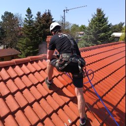 REGATECH FIRMA HANDLOWO-USŁUGOWA MIROSŁAW PRZYMUSZAŁA - Pracownik w uprzęży, z tyłu, czyści czerwoną dachówkę za pomocą myjki ciśnieniowej, w tle drzewa i antena.