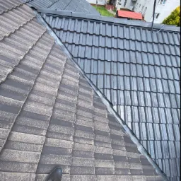 Częściowo wyczyszczony dach z szarej dachówki, widoczna różnica w kolorze i zabrudzeniach, noga w sportowym bucie na tle brudnej części dachu.