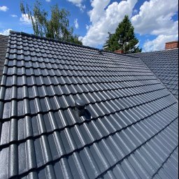 REGATECH FIRMA HANDLOWO-USŁUGOWA MIROSŁAW PRZYMUSZAŁA - Nowo pomalowany, lśniący, grafitowy dach z dachówki ceramicznej w słoneczny dzień, z widocznym kominkiem wentylacyjnym i fragmentem komina z cegły w tle, na tle błękitnego nieba z białymi obłokami.