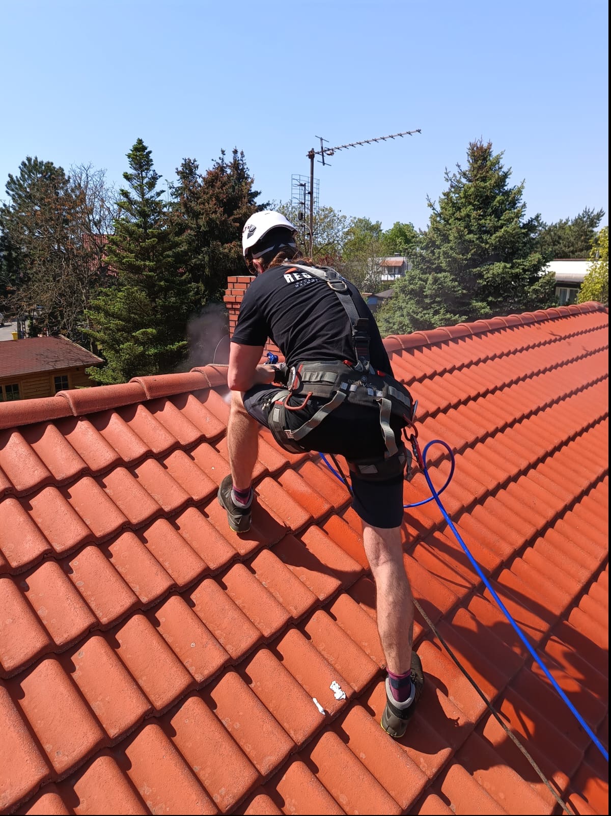 Pracownik w uprzęży, z tyłu, czyści czerwoną dachówkę za pomocą myjki ciśnieniowej, w tle drzewa i antena.