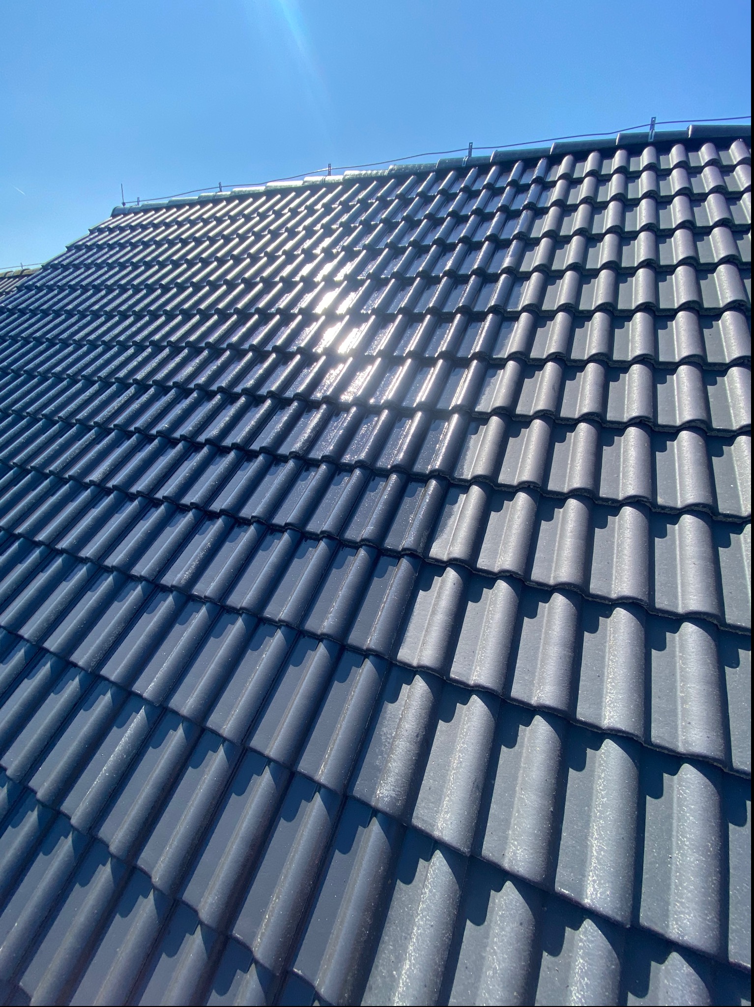 Błyszczący, grafitowy dach z dachówki ceramicznej w promieniach słońca, widok z dołu na tle błękitnego nieba.