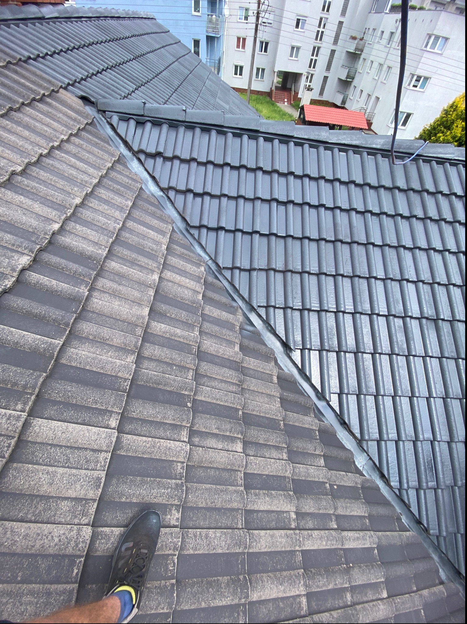 Częściowo wyczyszczony dach z szarej dachówki, widoczna różnica w kolorze i zabrudzeniach, noga w sportowym bucie na tle brudnej części dachu.