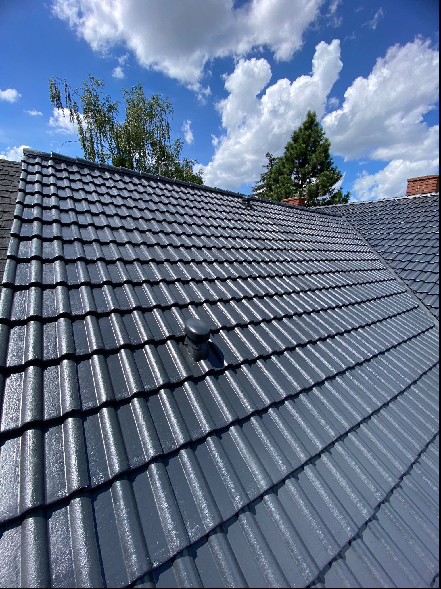 Nowo pomalowany, lśniący, grafitowy dach z dachówki ceramicznej w słoneczny dzień, z widocznym kominkiem wentylacyjnym i fragmentem komina z cegły w tle, na tle błękitnego nieba z białymi obłokami.