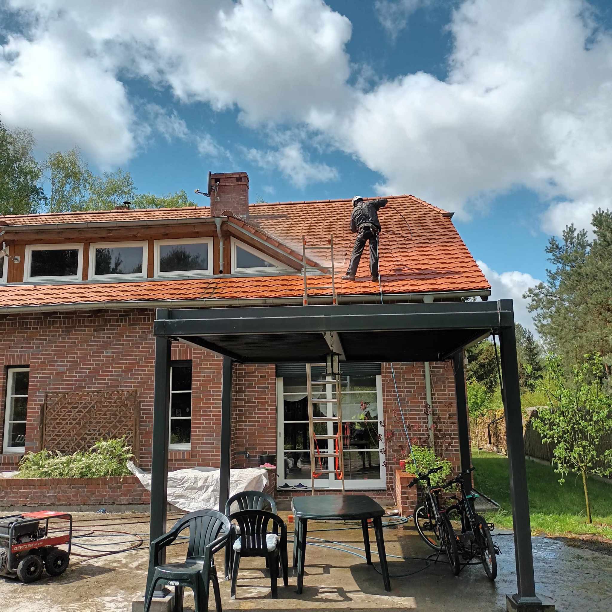 Pracownik w kombinezonie i kasku zabezpieczającym czyści dach z czerwonej dachówki za pomocą myjki ciśnieniowej; w tle ceglany dom z oknami, pergola, generator i błękitne niebo z chmurami.