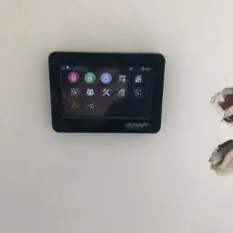 Czarny panel dotykowy systemu alarmowego marki Ropam zamontowany na białej ścianie, widoczne ikony funkcji i uszkodzenia tynku obok.