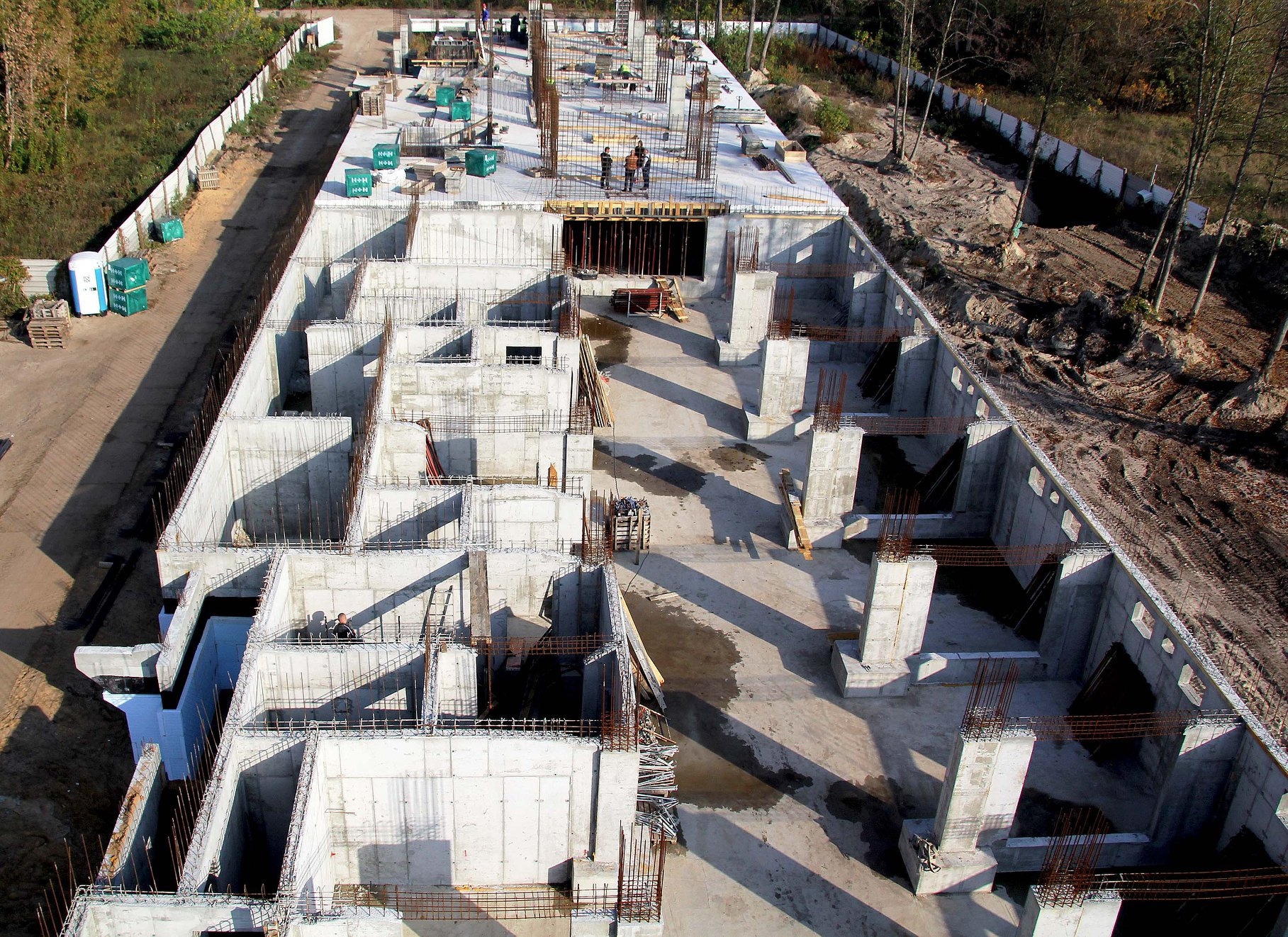 Widok z góry na budowę wielokondygnacyjnego budynku z betonu, z widocznymi zbrojeniami i trzema pracownikami na stropie, otoczonego drzewami.