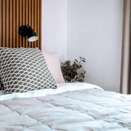 Yevhenii Honcharuk - Jasna sypialnia z drewnianym zagłówkiem, wzorzystymi poduszkami i lampką nocną, widok na balkon przez okno z beżowymi zasłonami.