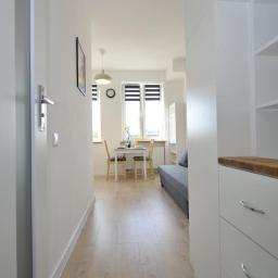 Yevhenii Honcharuk - Jasne wnętrze mieszkania po remoncie: widok z korytarza na salon z jadalnią, białe ściany, drewniana podłoga, stół z nakryciem, szara sofa, zabudowa kuchenna z drewnianym blatem.