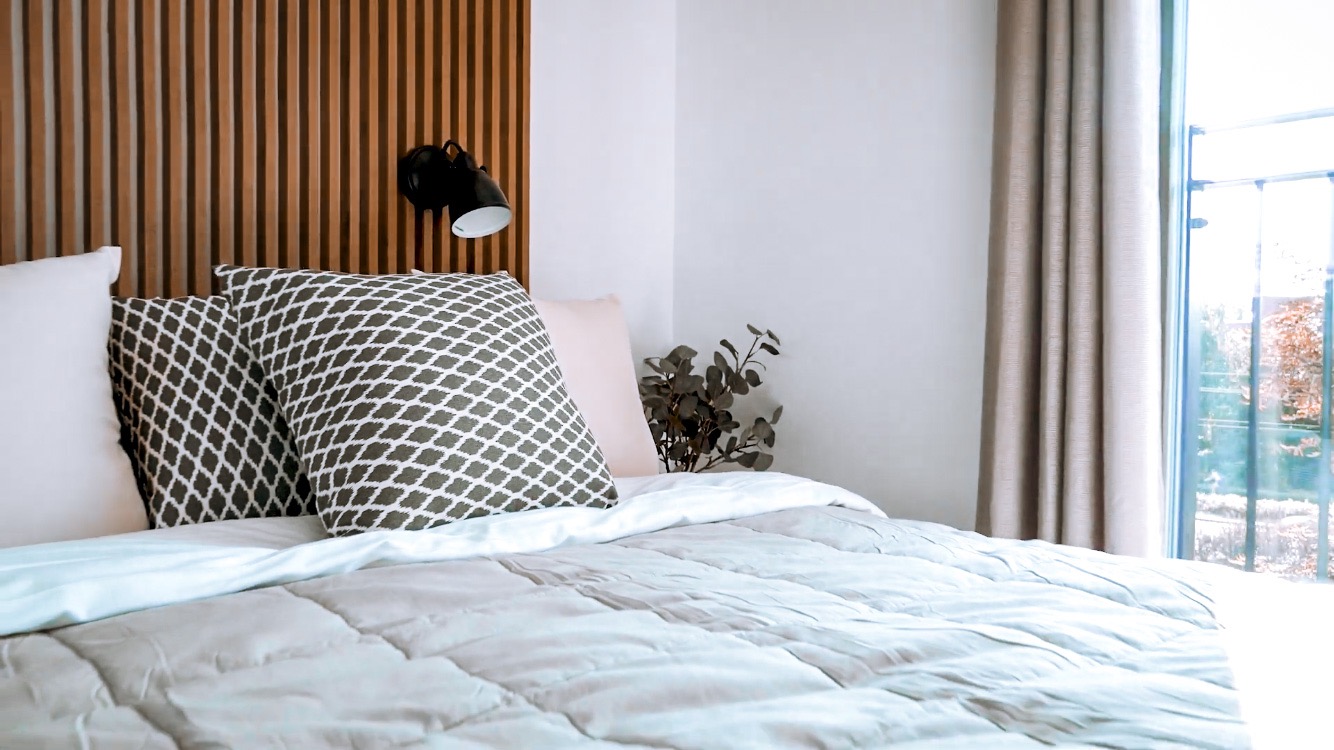 Jasna sypialnia z drewnianym zagłówkiem, wzorzystymi poduszkami i lampką nocną, widok na balkon przez okno z beżowymi zasłonami.