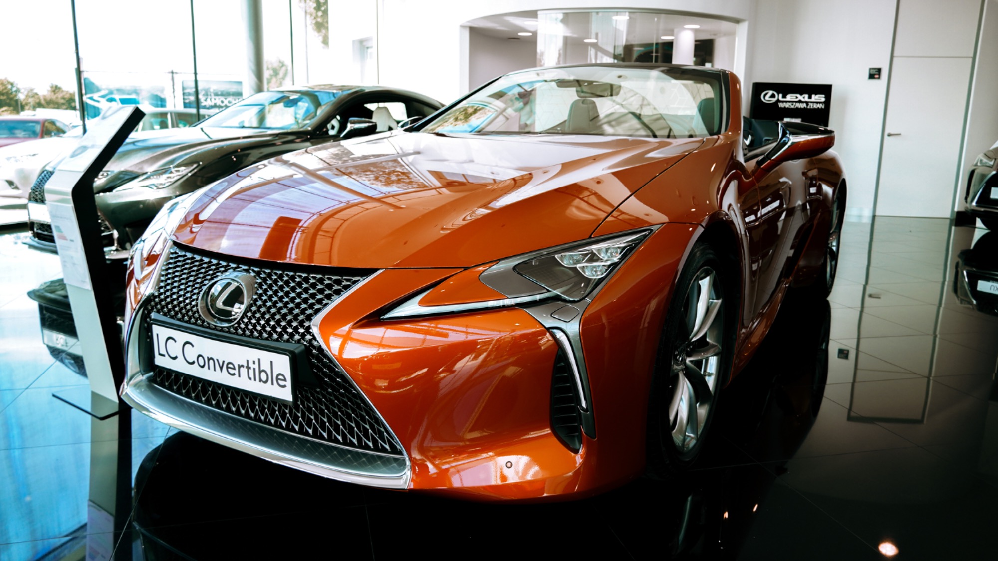 Pomarańczowy Lexus LC Convertible na błyszczącej podłodze salonu samochodowego, widoczny znak Lexus Warszawa Żerań w tle.