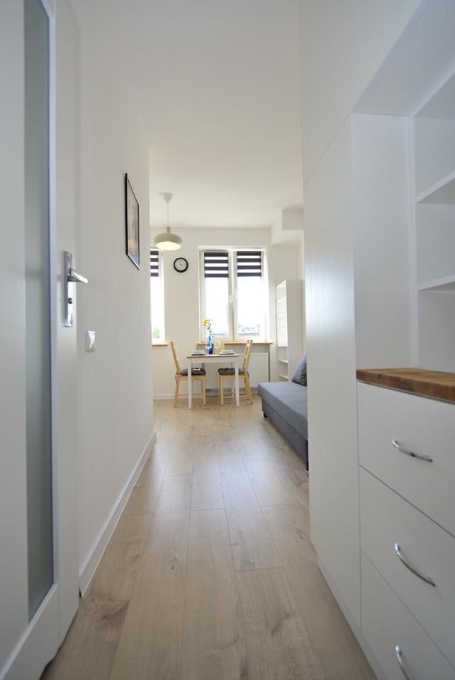 Jasne wnętrze mieszkania po remoncie: widok z korytarza na salon z jadalnią, białe ściany, drewniana podłoga, stół z nakryciem, szara sofa, zabudowa kuchenna z drewnianym blatem.