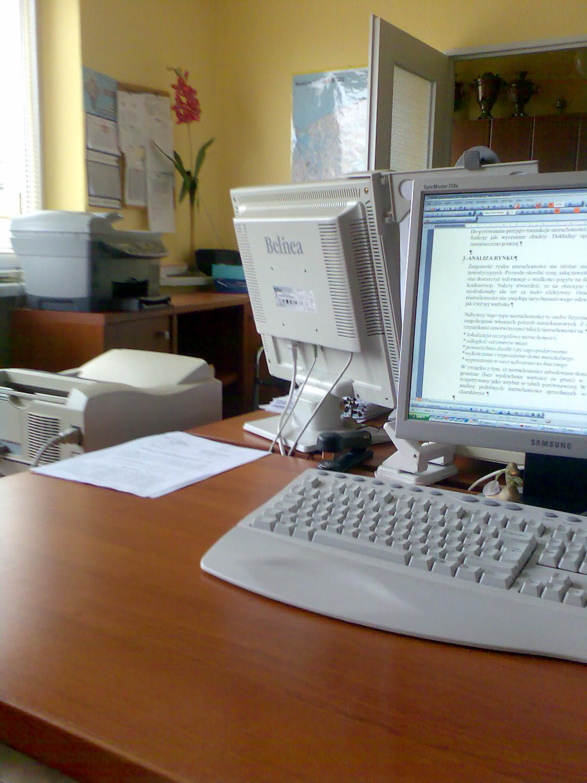 Biuro rzeczoznawcy: biurko z komputerami, klawiaturą, dokumentami, drukarką i kwiatem w tle, sugerujące pracę związaną z analizą rynku nieruchomości.