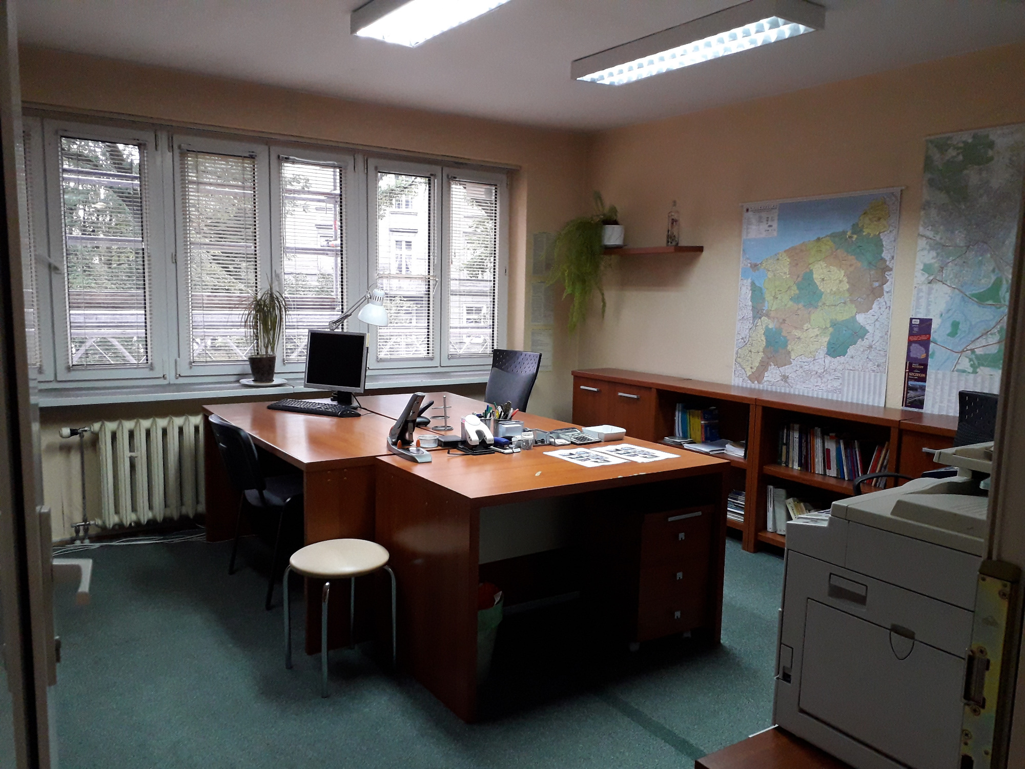 Biuro z dużym drewnianym biurkiem, komputerem, lampką, krzesłem, taboretem, drukarką, regałem z książkami i mapami na ścianie.