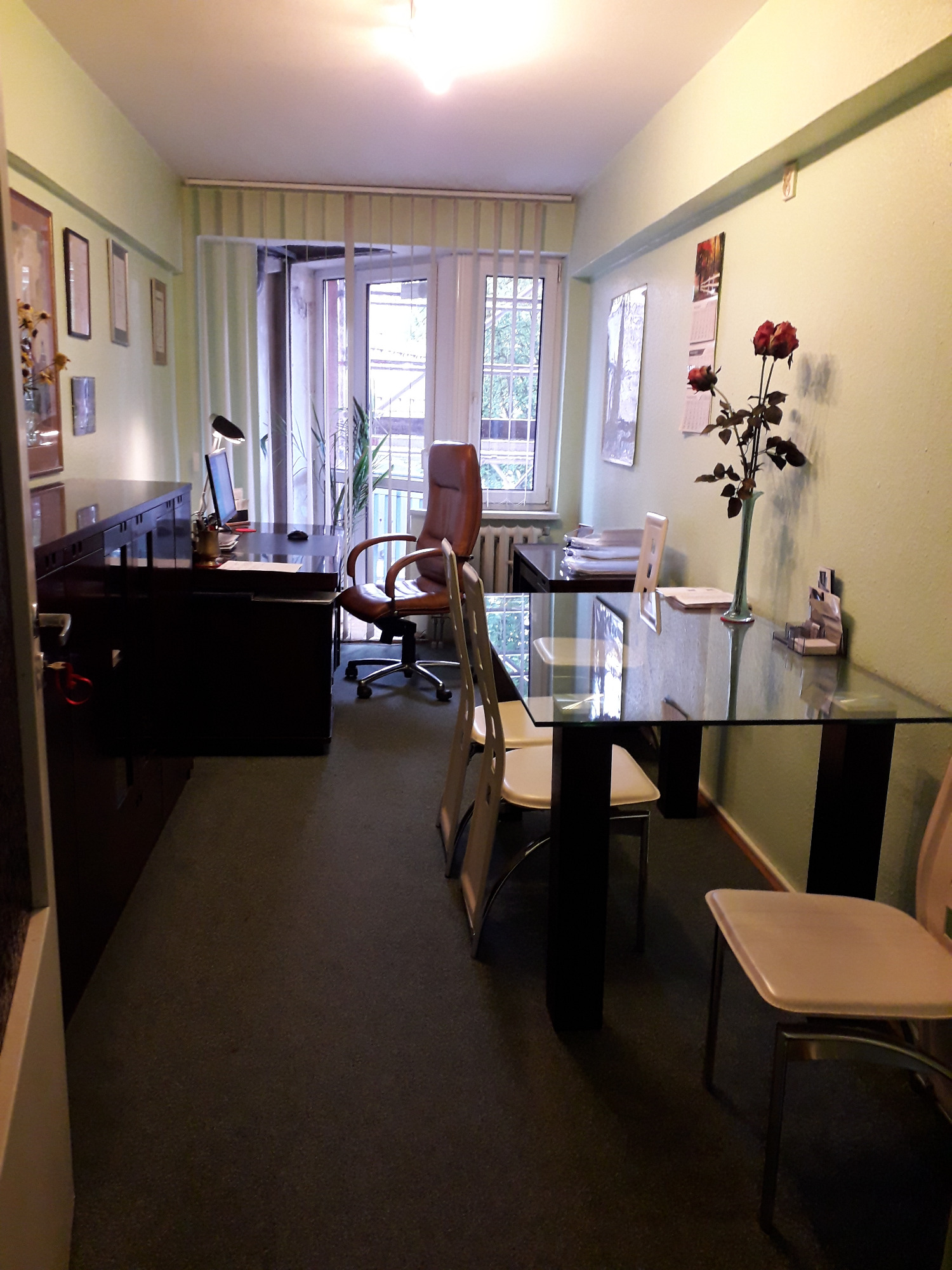 Biuro z ciemnymi meblami, skórzanym fotelem obrotowym, szklanym stołem i trzema czerwonymi różami w wazonie na ścianie w tle kalendarz.