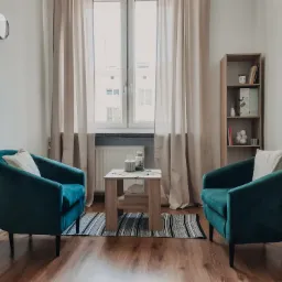 Gabinet terapeutyczny z dwoma turkusowymi fotelami, stolikiem kawowym z dekoracjami i oknem z zasłonami w tle.