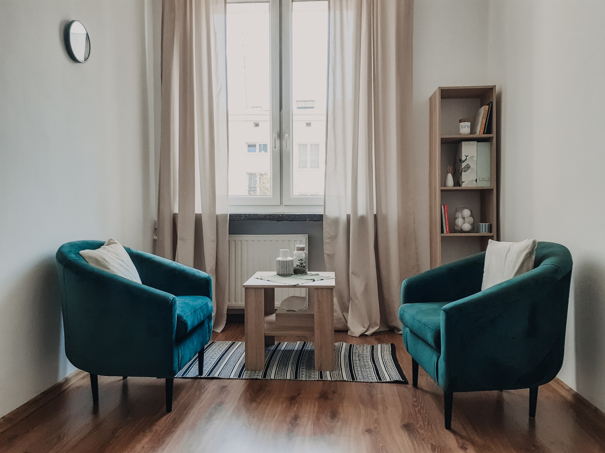 Gabinet terapeutyczny z dwoma turkusowymi fotelami, stolikiem kawowym z dekoracjami i oknem z zasłonami w tle.