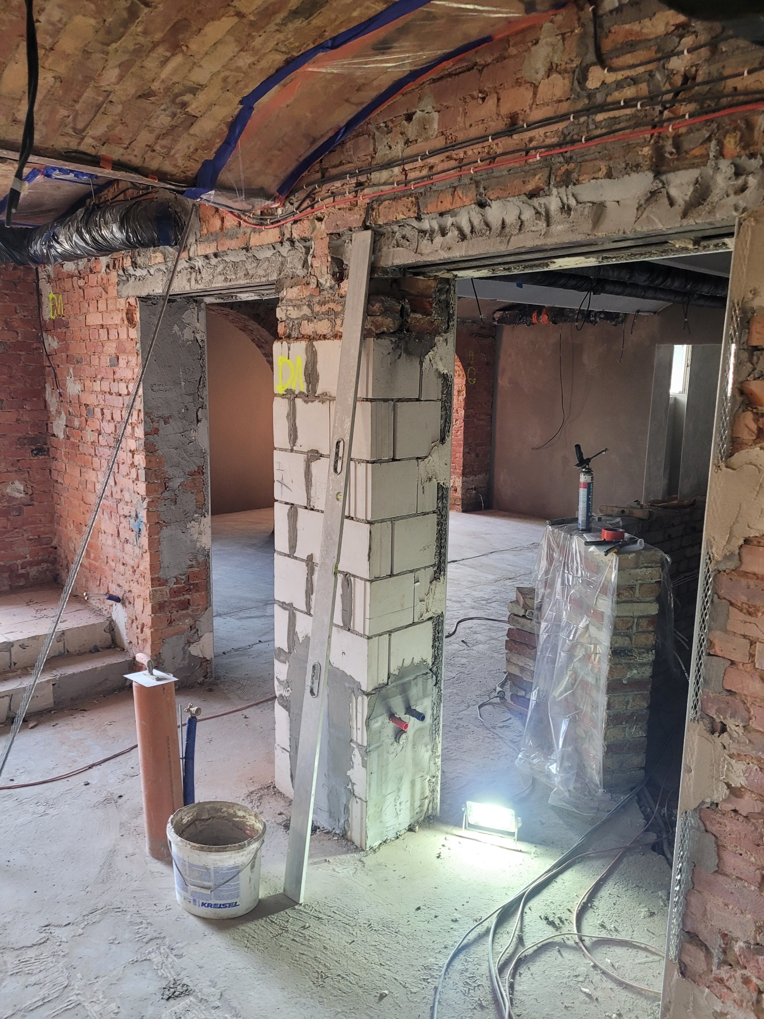 Wnętrze w trakcie remontu: surowa cegła, instalacje na suficie, wznoszone ściany z bloczków, poziomica oparta o ścianę, wiadro z zaprawą Kreisel, oświetlenie robocze na podłodze.