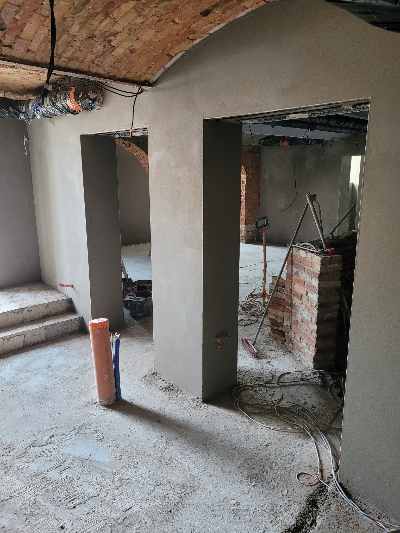 Wnętrze w trakcie tynkowania maszynowego; widoczne gładkie, szare ściany, odsłonięta cegła w łukowym przejściu, narzędzia i kable na podłodze, oraz fragment instalacji wentylacyjnej pod ceglanym...