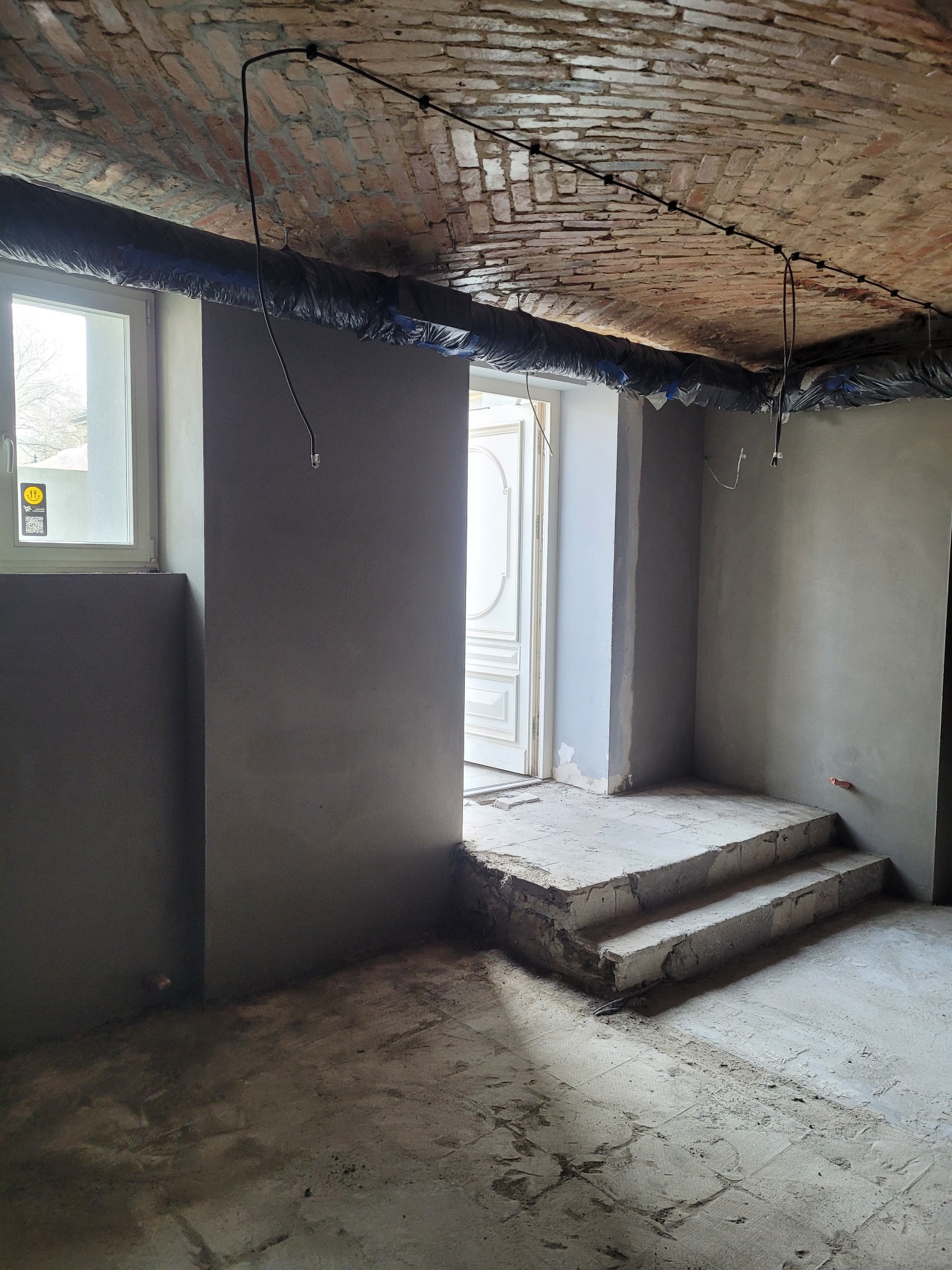 Wnętrze w trakcie prac wykończeniowych: ściany pokryte świeżym, szarym tynkiem, widoczne instalacje elektryczne pod sufitem z cegły, zabezpieczone folią, oraz schody z surowego betonu prowadzące...