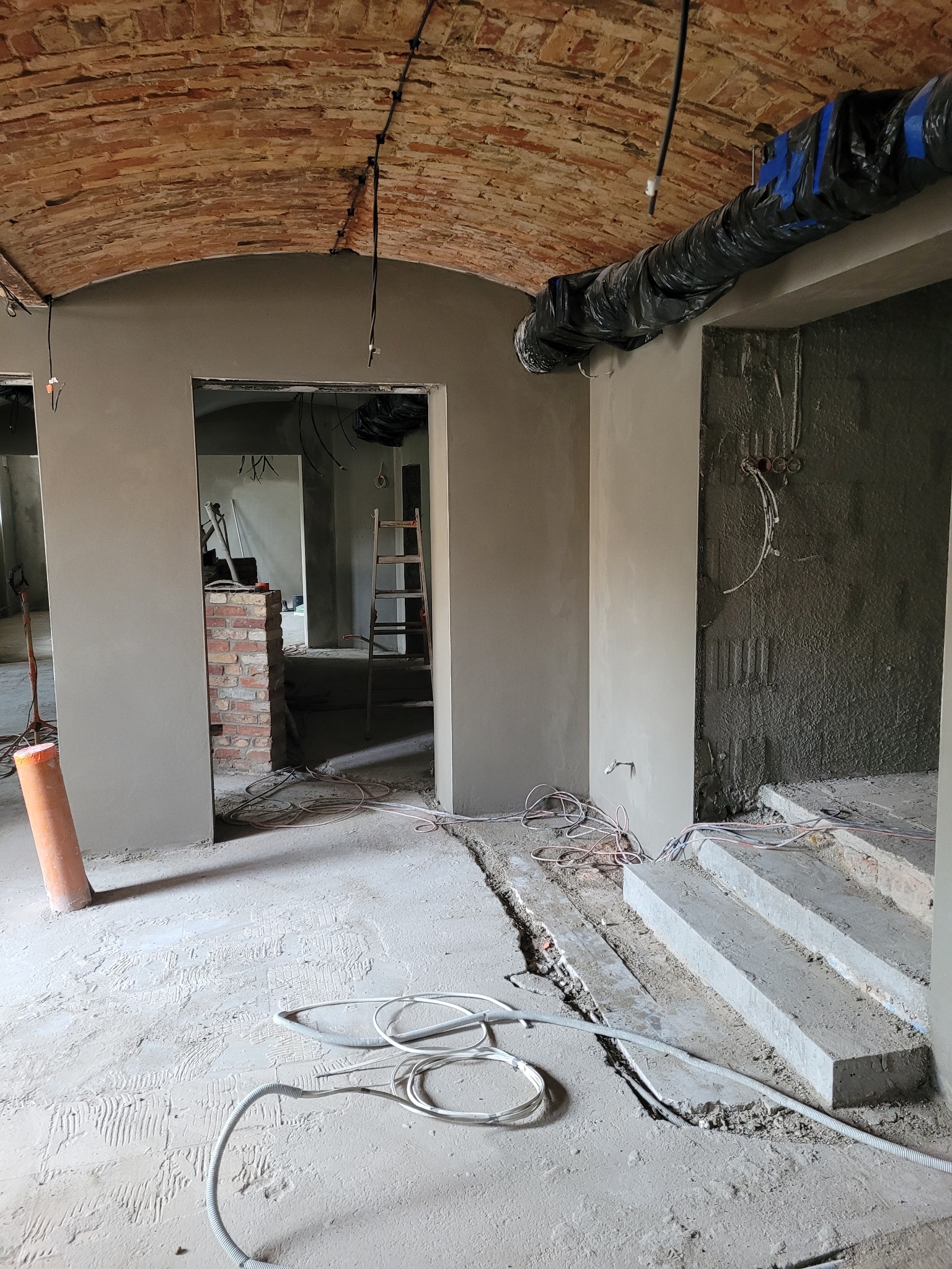 Wnętrze w trakcie prac wykończeniowych: ściany pokryte świeżym tynkiem, widoczny surowy ceglany łuk, wystające przewody elektryczne i rury wentylacyjne, na podłodze resztki materiałów budowlanych...