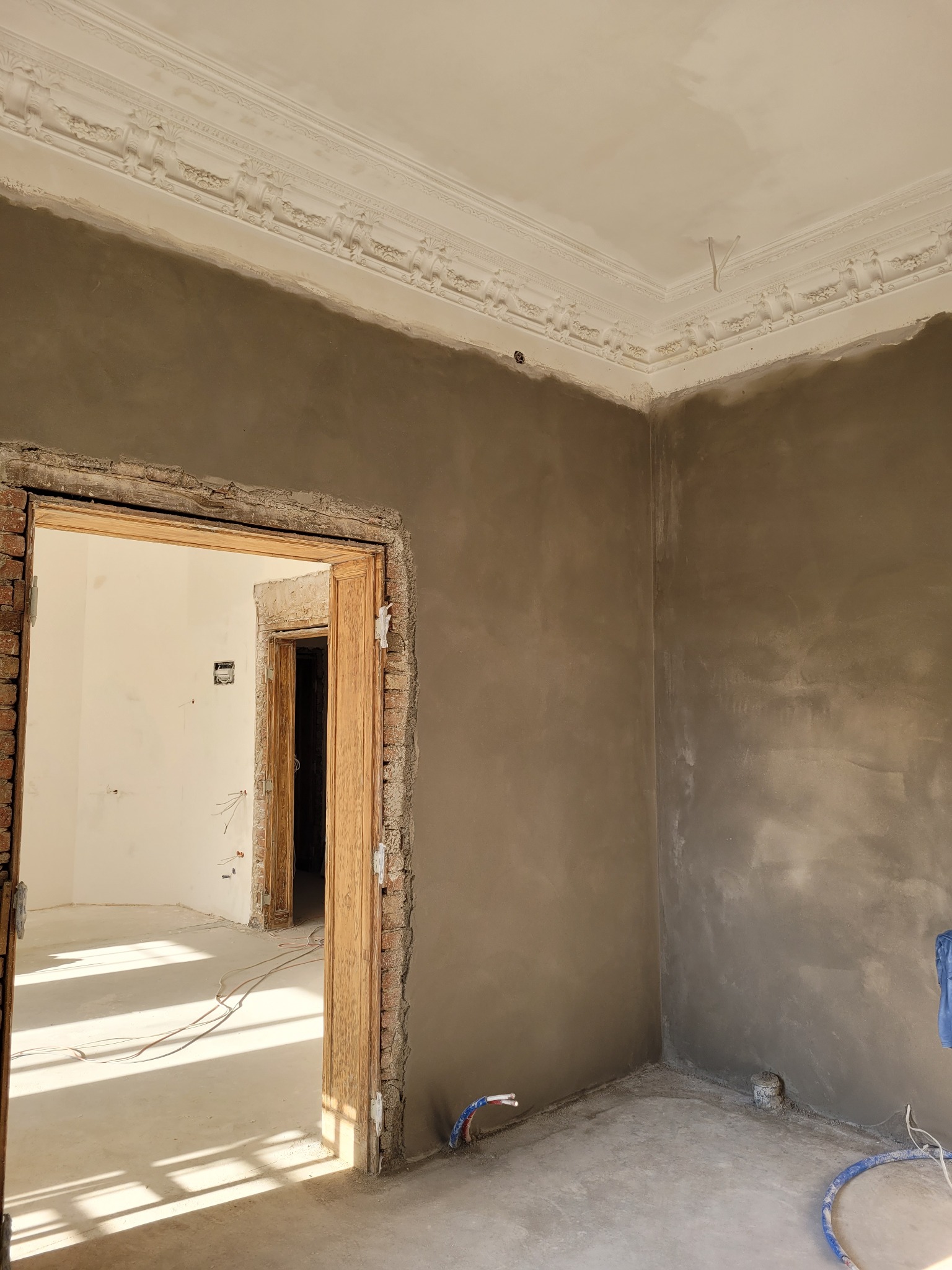 Pokój w trakcie prac. Ściany pokryte świeżym, szarym tynkiem, widoczna surowa cegła wokół drewnianych futryn drzwi, dekoracyjny gzyms sufitowy.