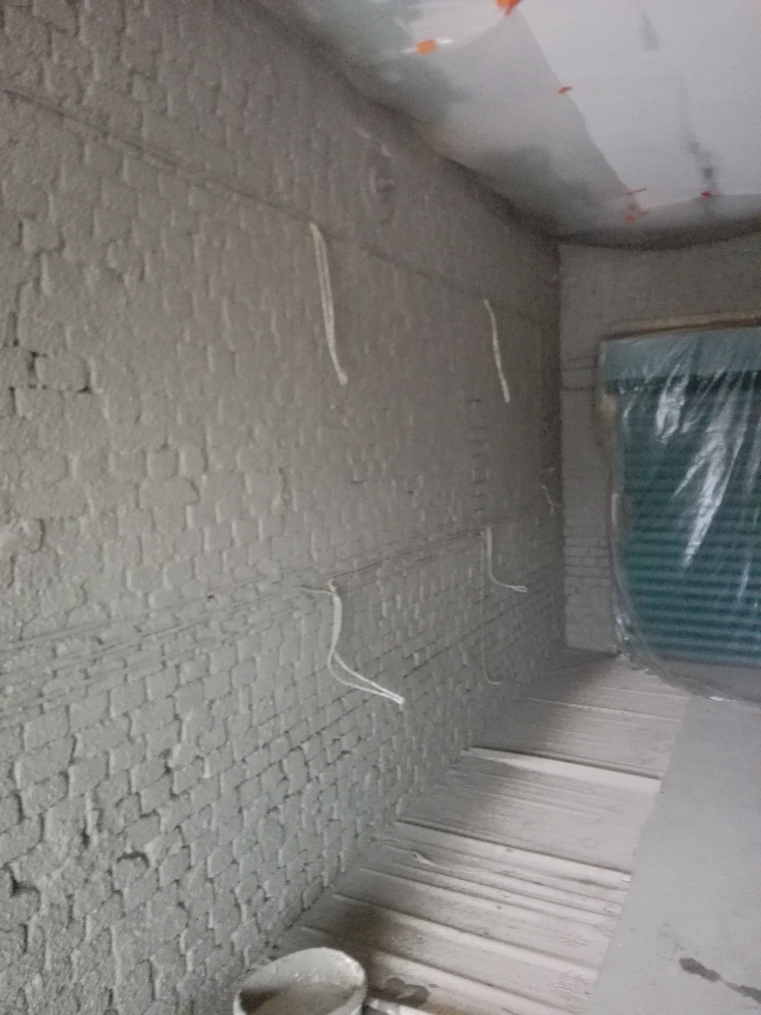 Świeżo nałożona, szara warstwa na murze z cegieł z widocznymi przewodami elektrycznymi, podłoga zabezpieczona deskami, okno zasłonięte folią ochronną.