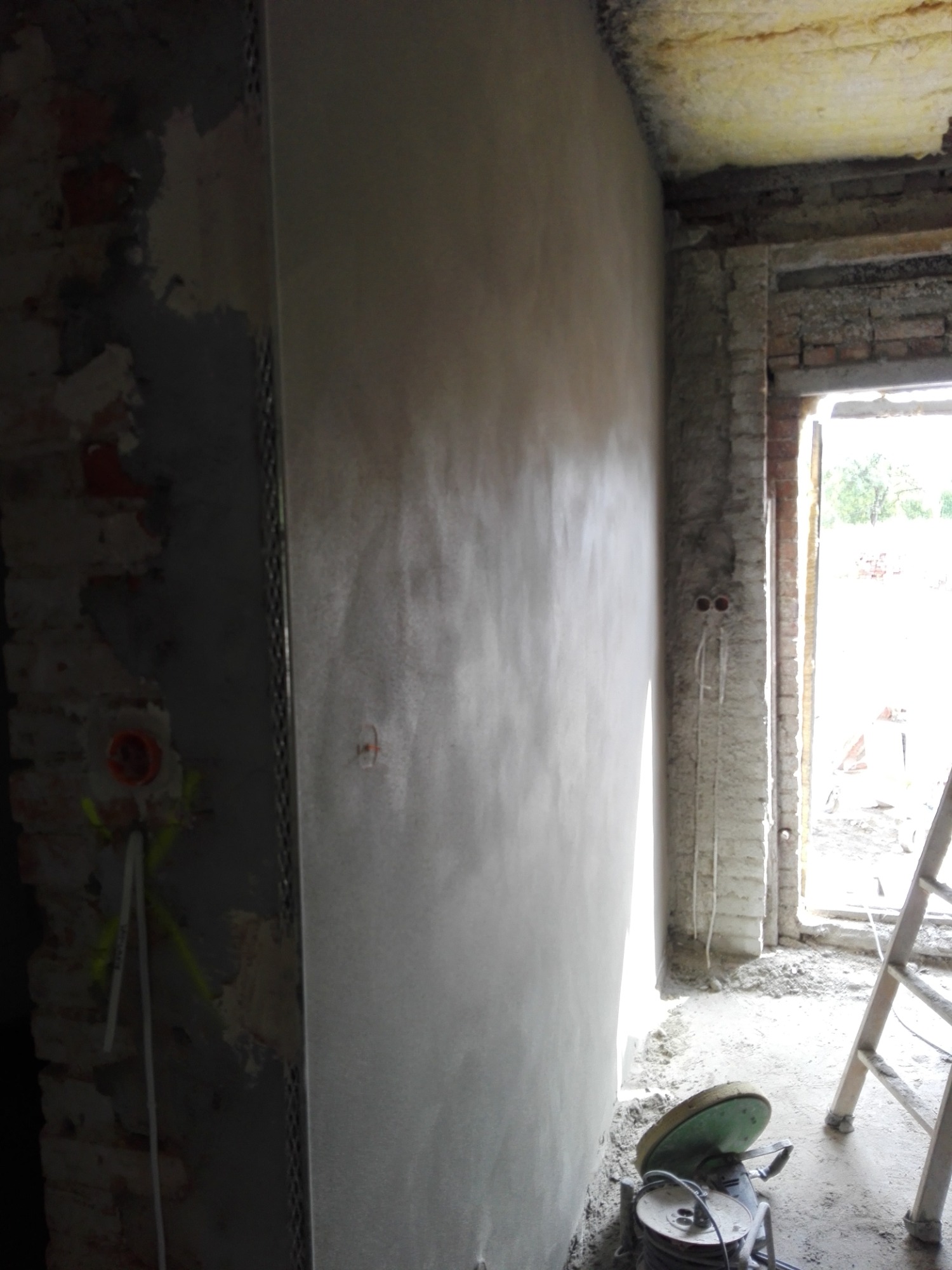 Świeżo nałożony, wilgotny tynk maszynowy na ścianie w surowym wnętrzu budynku, widoczne przewody elektryczne i drabina w tle.