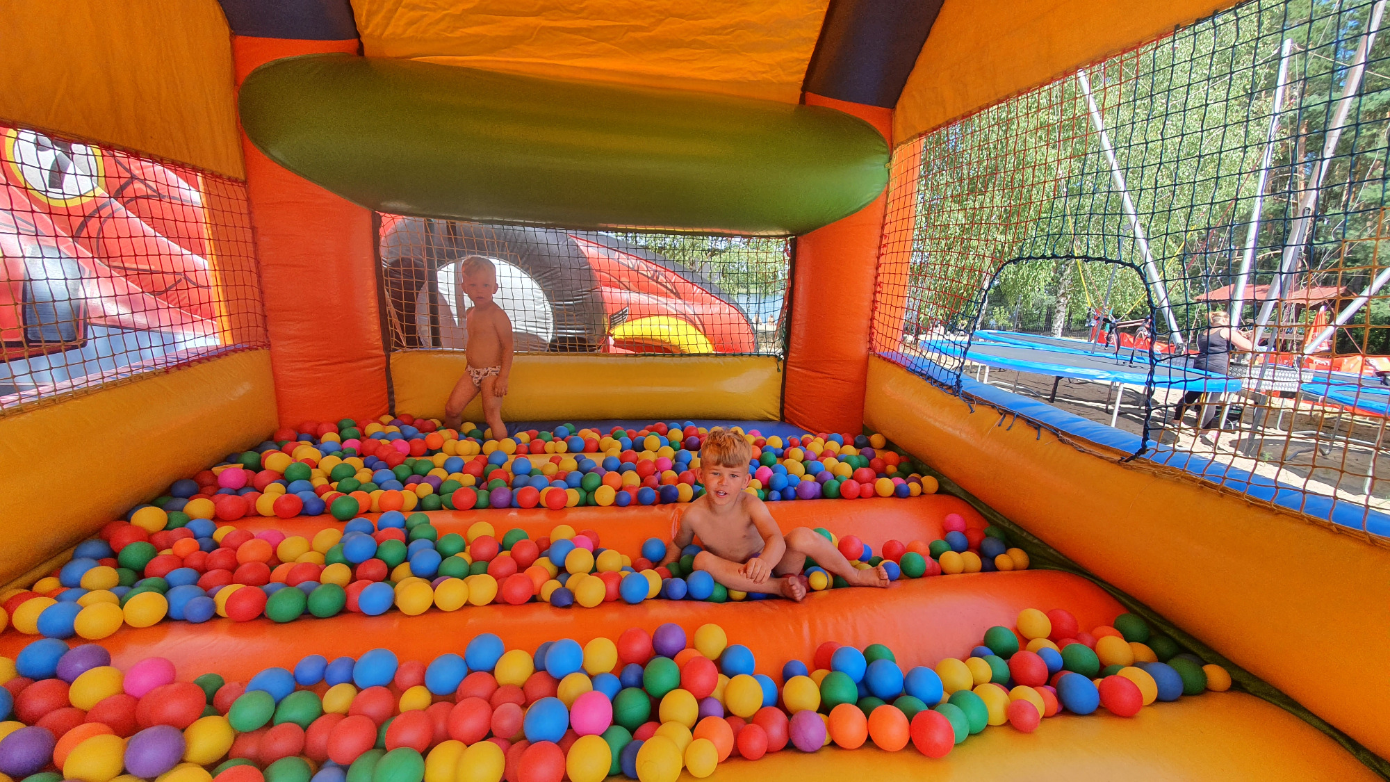 Dzieci bawią się w pomarańczowo-żółtym dmuchanym zamku z kolorowymi piłeczkami w Osmolinie. W tle widoczny trampolinowy park rozrywki przez siatkę ochronną.