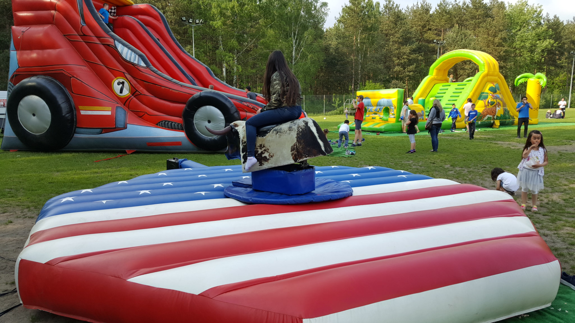 Dziewczyna siedzi na mechanicznym byku rodeo umieszczonym na dmuchanej platformie w barwach flagi USA. W tle dmuchane zamki i zjeżdżalnie oraz bawiące się dzieci.