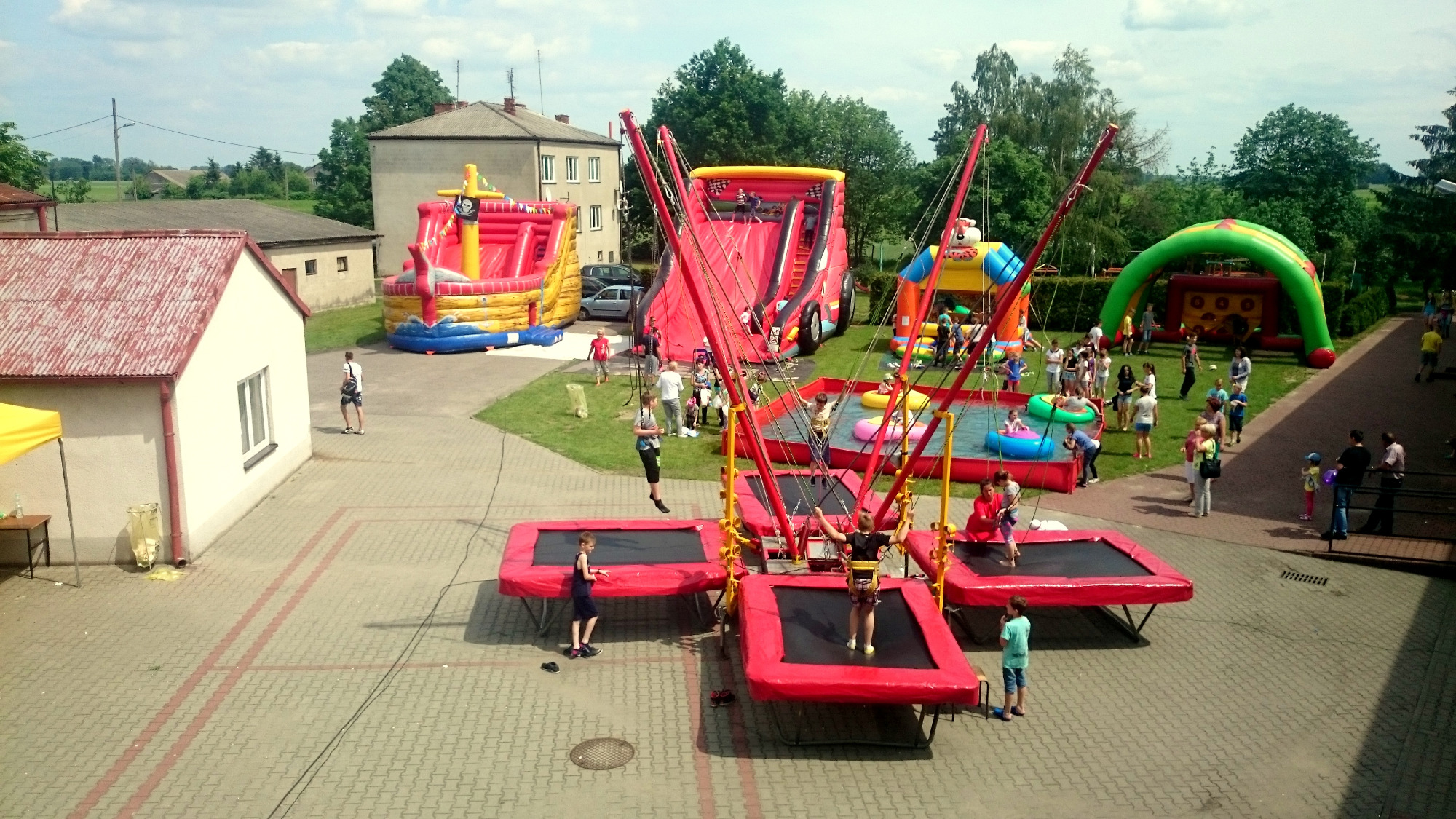Zewnętrzna strefa zabaw z dmuchanymi zamkami, zjeżdżalniami i trampolinami bungee, na których dzieci skaczą w uprzężach, otoczona zielenią i budynkami.