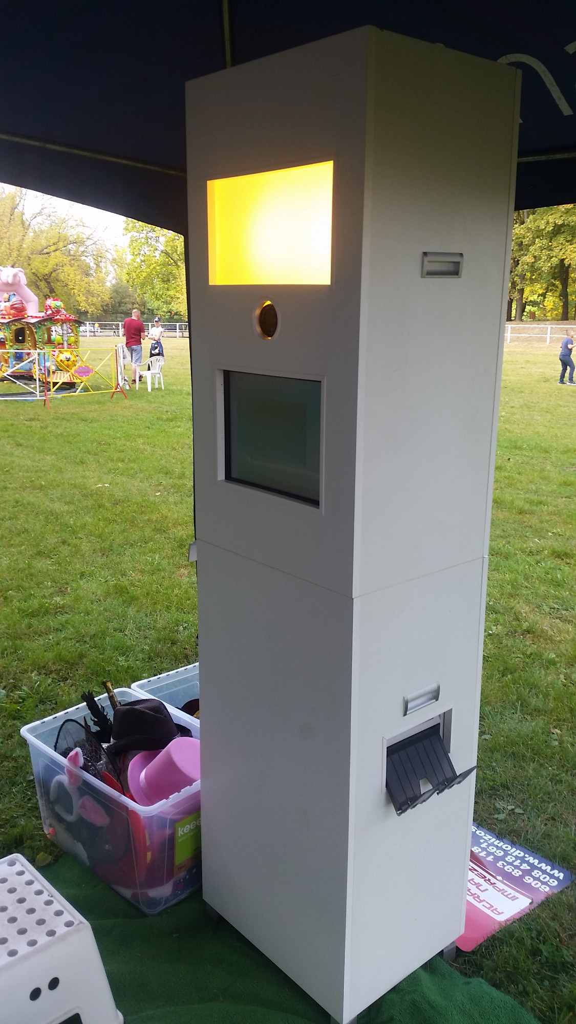Biała budka fotograficzna z żółtym podświetleniem ekranu, obok pojemnik z rekwizytami imprezowymi na trawie, w tle karuzela i ludzie.