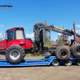 SecureTrans - Czerwony harwester Valmet 890.3 z chwytakiem, transportowany na niskopodwoziowej, niebieskiej lawecie VS-mont, na tle polnej drogi i błękitnego nieba.